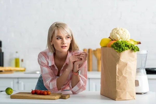 Femme blonde rêveuse près de sac en papier avec épicerie dans la cuisine — Photo de stock