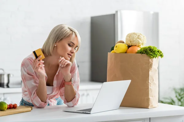 Menina loira atraente segurando cartão de crédito enquanto olha para laptop perto de saco de papel com mantimentos — Fotografia de Stock