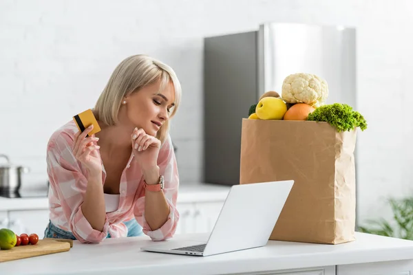 Attraente ragazza bionda in possesso di carta di credito mentre guardando il computer portatile vicino sacchetto di carta con generi alimentari — Foto stock