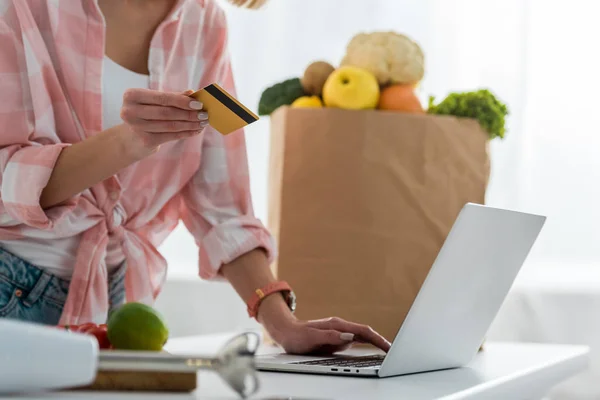 Vue recadrée de la jeune femme tenant la carte de crédit tout en utilisant un ordinateur portable près du sac en papier avec épicerie — Photo de stock