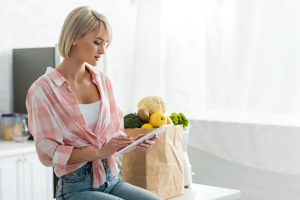 Atractiva mujer rubia usando tableta digital cerca de bolsa de papel con ingredientes - foto de stock