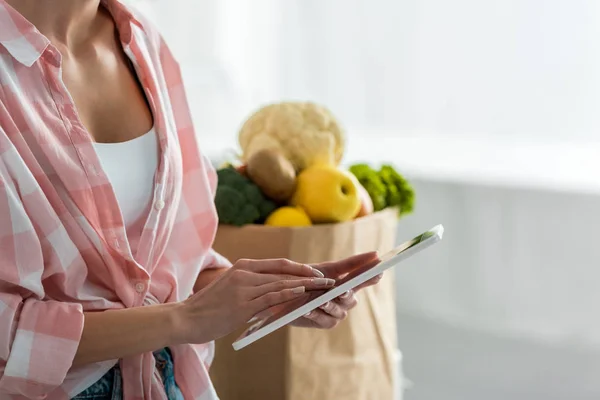 Vista ritagliata della donna utilizzando tablet digitale vicino sacchetto di carta con ingredienti — Stock Photo