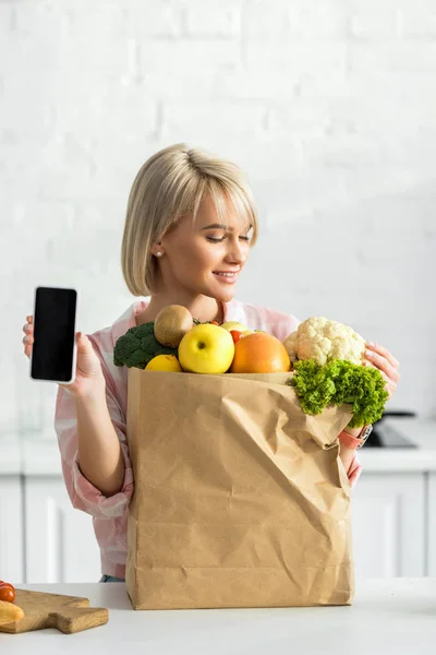 Весела блондинка тримає смартфон з порожнім екраном біля паперового мішка з інгредієнтами — стокове фото