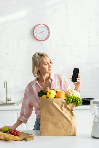 Mujer rubia feliz sosteniendo teléfono inteligente con pantalla en blanco cerca de bolsa de papel con ingredientes - foto de stock