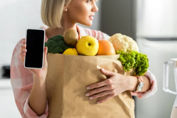 Vista ritagliata della ragazza bionda che tiene smartphone con schermo bianco vicino al sacchetto di carta con generi alimentari — Foto stock