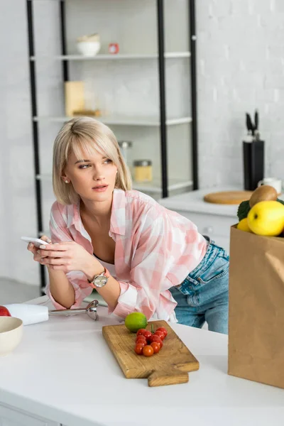 Chica rubia usando smartphone cerca de bolsa de papel con comestibles - foto de stock