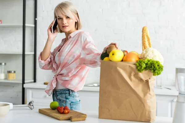 Attractive blonde girl talking on smartphone near paper bag with groceries — Stock Photo