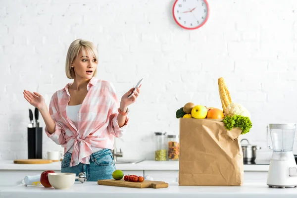 Chica rubia sorprendida con teléfono inteligente cerca de bolsa de papel con comestibles - foto de stock