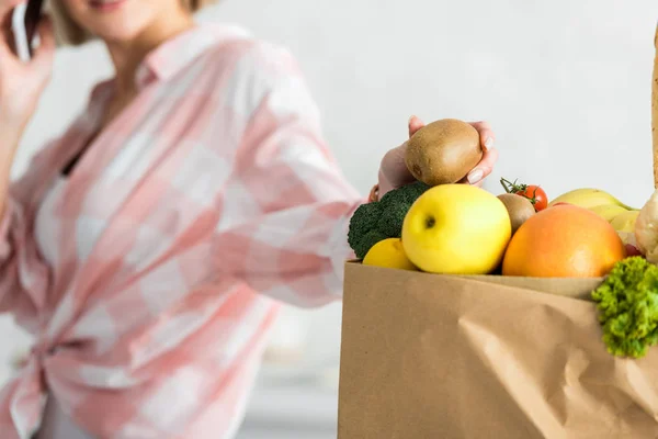 Cropped view of woman holding kiwi fruit near paper bag with food — Stock Photo