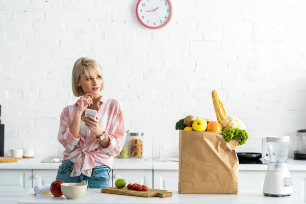 Chica rubia pensativa con teléfono inteligente cerca de bolsa de papel con comestibles - foto de stock