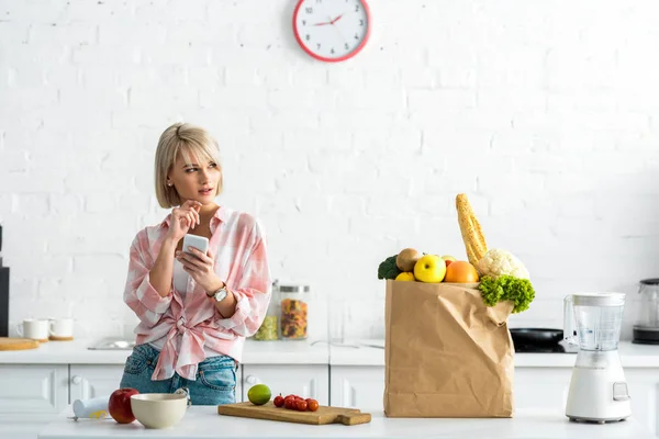 Chère fille blonde avec smartphone près du sac en papier avec épicerie — Photo de stock