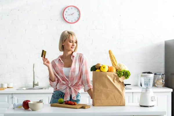 Atractiva chica rubia sosteniendo tarjeta de crédito cerca de bolsa de papel con comestibles - foto de stock