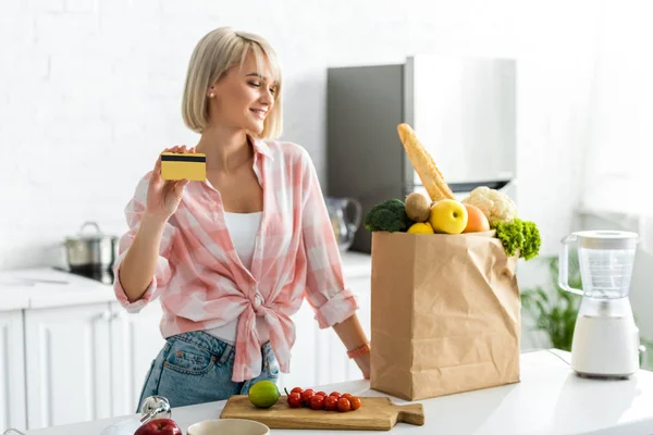 Жизнерадостная блондинка держит кредитку рядом с бумажным пакетом с продуктами — стоковое фото