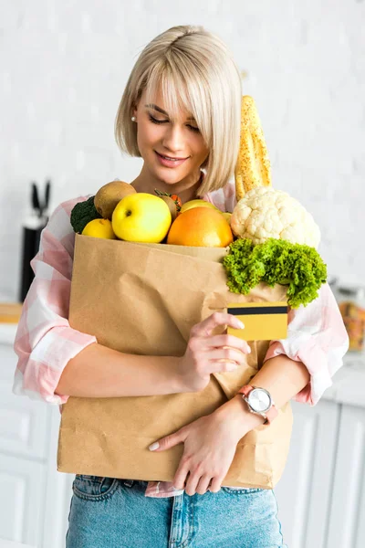 Chica rubia feliz sosteniendo tarjeta de crédito mientras abraza bolsa de papel con comestibles - foto de stock