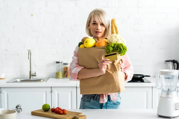 Surprised blonde girl holding credit card near paper bag with groceries — Stock Photo