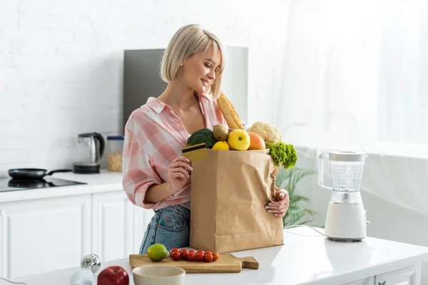 Feliz rubia joven sosteniendo tarjeta de crédito cerca de bolsa de papel con comestibles - foto de stock