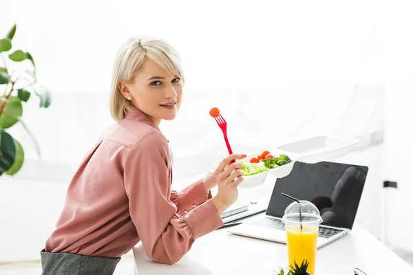 Freelancer loira alegre perto de laptop com tela em branco e legumes na caixa takeaway — Fotografia de Stock