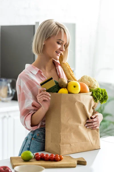 Mujer rubia feliz sosteniendo la tarjeta de crédito cerca de bolsa de papel con comestibles - foto de stock