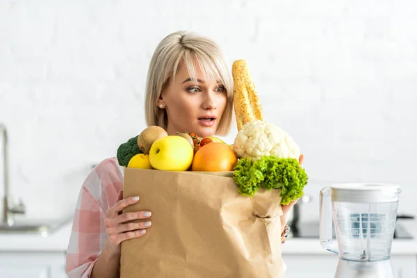 Überrascht blonde junge Frau umarmt Papiertüte mit Lebensmitteln und blickt auf Mixer — Stockfoto