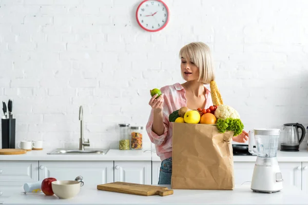 Mujer rubia sorprendida sosteniendo cal cerca de bolsa de papel con comestibles - foto de stock