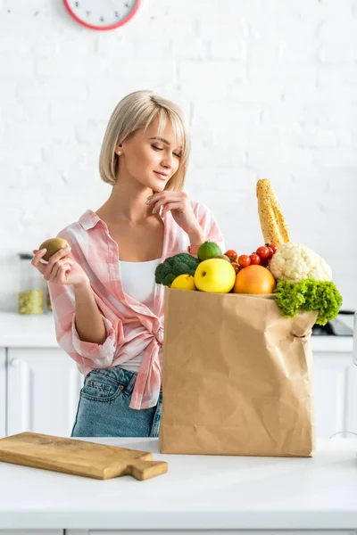 Happy blonde young woman holding kiwi fruit near paper bag with groceries — Stock Photo