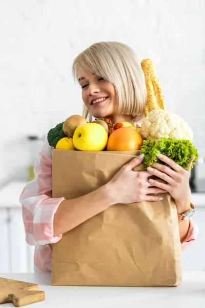 Feliz rubia joven mujer abrazando bolsa de papel con comestibles - foto de stock