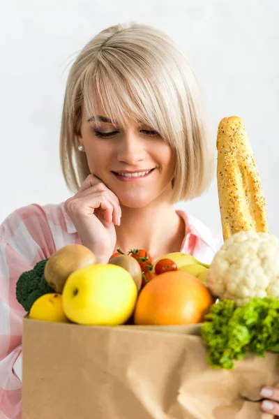 Glückliche blonde junge Frau schaut auf Papiertüte mit Lebensmitteln — Stockfoto