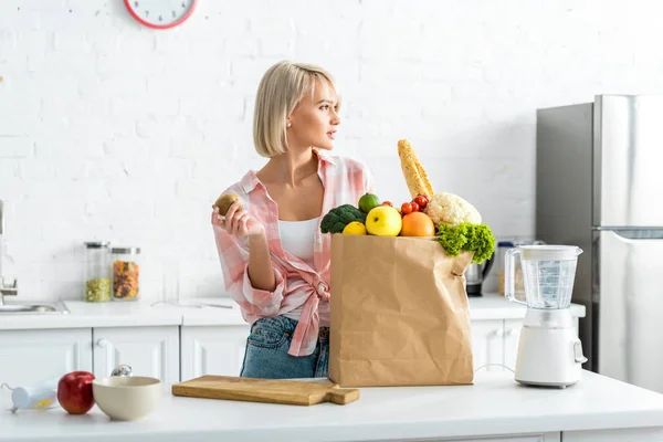 Приваблива блондинка тримає ківі фрукти біля паперового мішка з продуктами — стокове фото
