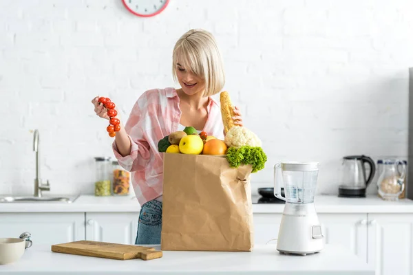 Glückliche blonde Frau blickt auf Kirschtomaten in der Nähe von Papiertüten mit Lebensmitteln — Stockfoto