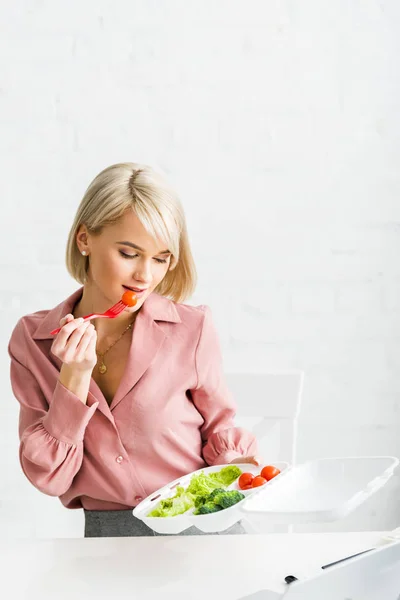 Menina loira comendo tomate cereja e olhando para caixa takeaway — Fotografia de Stock