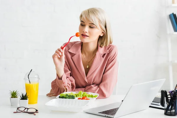 Freelancer rubia sosteniendo tenedor con tomate cereza cerca del portátil - foto de stock