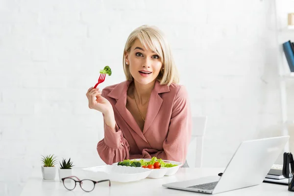 Sorprendida freelancer rubia sosteniendo tenedor con tomate cereza cerca del ordenador portátil - foto de stock