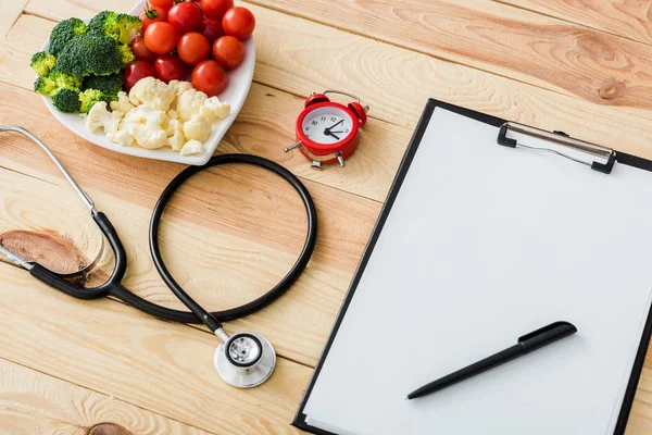 Presse-papiers vierge avec stylo près du stéthoscope, réveil et légumes savoureux sur plaque en forme de cœur — Photo de stock