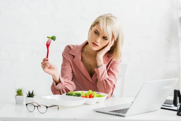 Upset freelancer holding fork with cherry tomato near laptop — Stock Photo