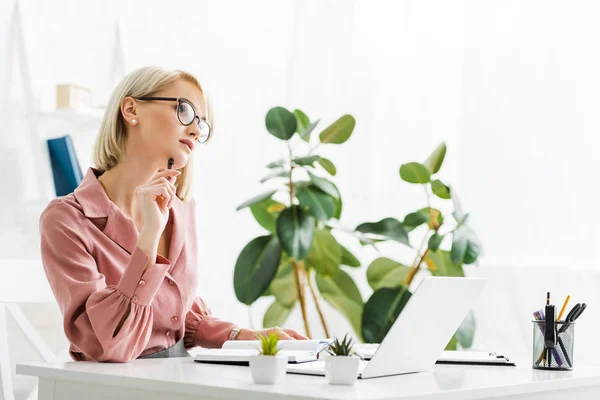 Pensieroso giovane bionda freelance in occhiali seduto vicino al computer portatile — Foto stock