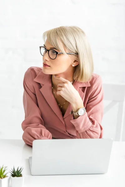 Nachdenkliche junge blonde Freiberuflerin mit Brille sitzt neben Laptop — Stockfoto
