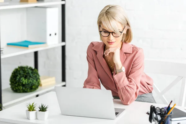 Nachdenkliche junge blonde Freiberuflerin mit Brille blickt auf Laptop — Stockfoto