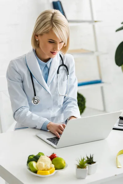 Nutricionista loira usando laptop perto da placa com alimentos orgânicos e saborosos — Stock Photo