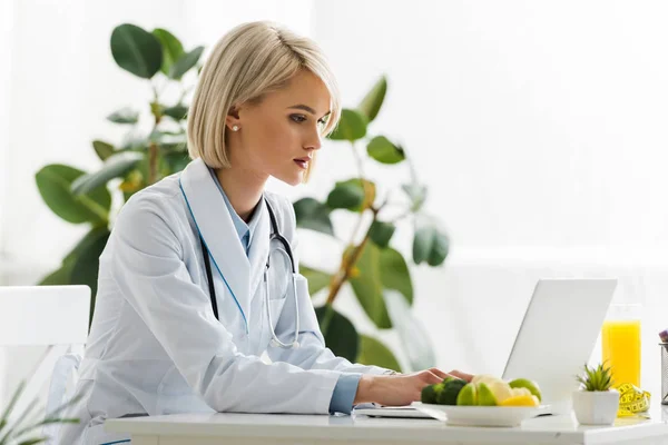 Beautiful blonde nutritionist using laptop near plate with organic food and glass of orange juice — Stock Photo