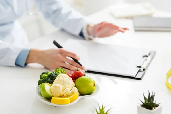 Abgeschnittene Ansicht eines Ernährungswissenschaftlers, der Stift in der Nähe von Klemmbrett und Teller mit Bio-Lebensmitteln hält — Stockfoto