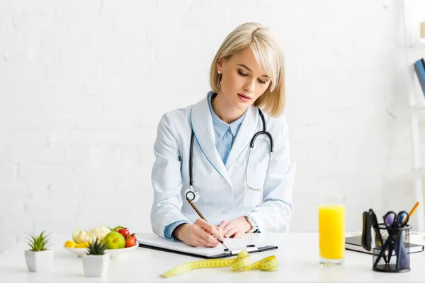 Attractive blonde nutritionist in white coat writing on clipboard — Stock Photo