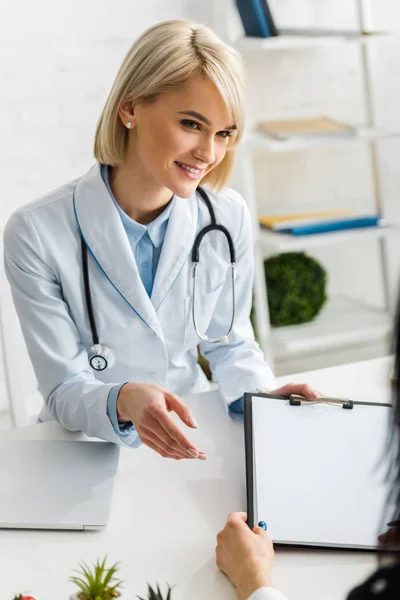 Fröhliche blonde Ernährungsberaterin im weißen Mantel übergibt leeres Klemmbrett an Patientin — Stockfoto