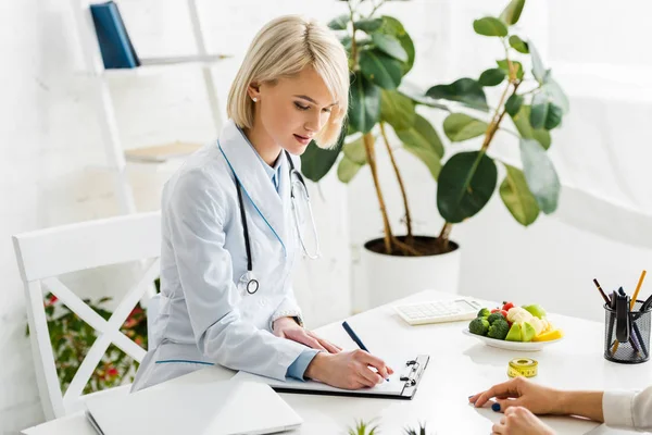 Atractiva rubia nutricionista en blanco abrigo escritura diagnóstico cerca paciente - foto de stock