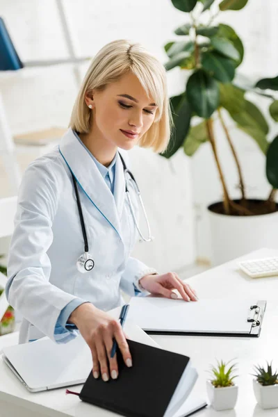 Attractive blonde doctor in white coat holding notebook near blank clipboard — Stock Photo