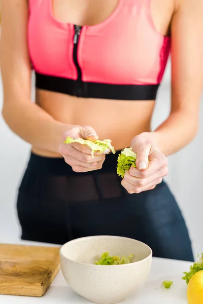 Cropped view of woman in sportswear holding salad leaves near bowl — Stock Photo