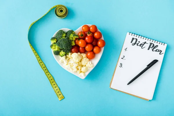 Vue de dessus des légumes savoureux sur la plaque en forme de coeur près du ruban à mesurer et du carnet avec lettrage de régime sur bleu — Photo de stock