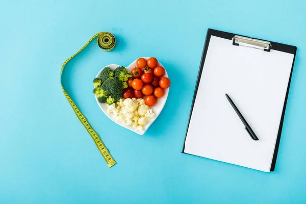 Top view of tasty vegetables on heart-shape plate near measuring tape, stethoscope and blank clipboard on blue — Stock Photo