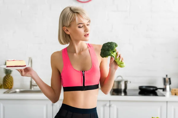 Blondes Mädchen blickt auf grünen Brokkoli und hält Untertasse mit süßem Kuchen in der Hand — Stockfoto
