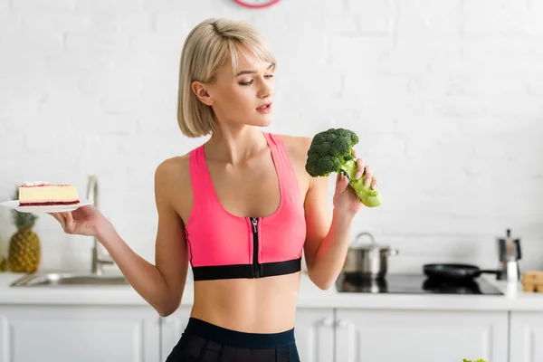 Blonde girl looking at green broccoli while holding saucer with sweet cake — Stock Photo