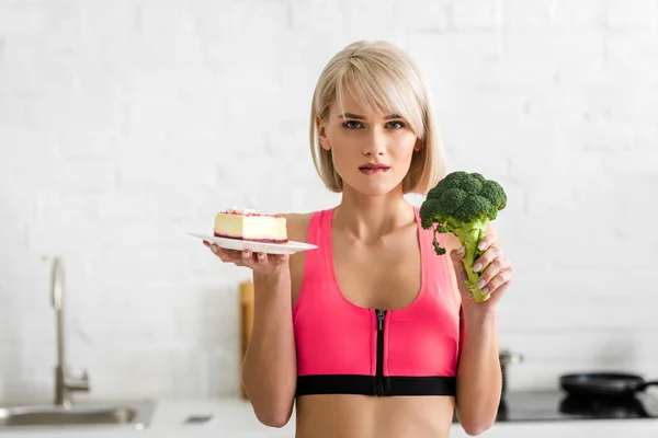 Blondes Mädchen hält grünen Brokkoli und Untertasse mit süßem Kuchen in der Hand und beißt Lippe — Stockfoto